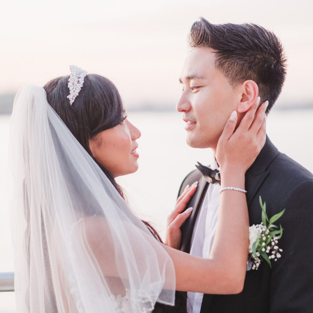 Bride touches groom's neck as they stand on a seafront promenade at sunset.
